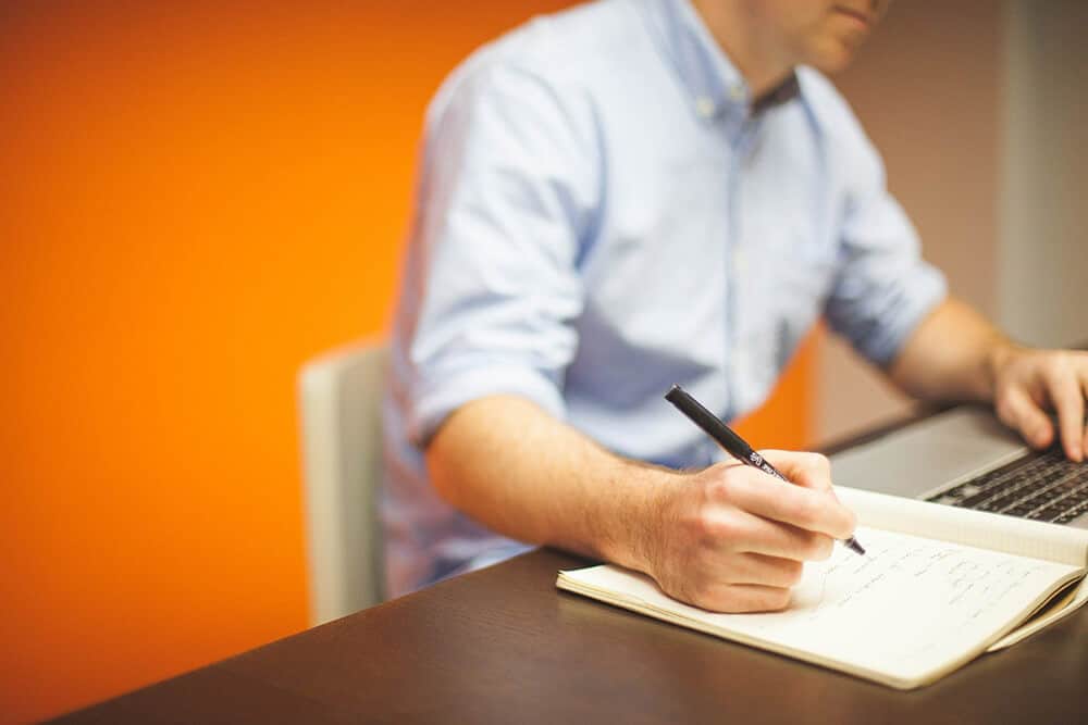 Person writing notes while using laptop